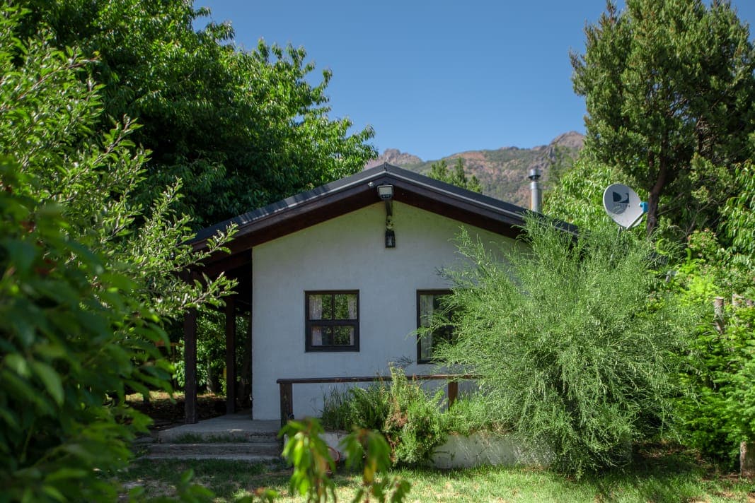 A one-story cabin is surrounded by lush greenery, featuring a simple exterior with a dark roof and white walls. Large windows allow natural light to enter, while a covered porch provides shade. Tall trees and shrubs create a natural and private setting.