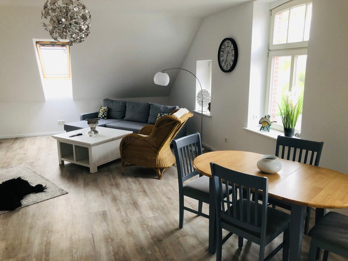 The living area features a comfortable gray sofa paired with a decorative chair. A wooden dining table is set for six, and a light rug defines the seating area. Natural light filters through large windows, complemented by contemporary lighting fixtures.