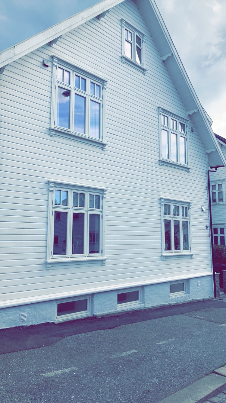 A well-maintained white wooden house features multiple large windows arranged symmetrically across the facade, allowing ample natural light. The sloping roof adds character, and the structure is set back slightly from the street, with a clear view of the surrounding area.