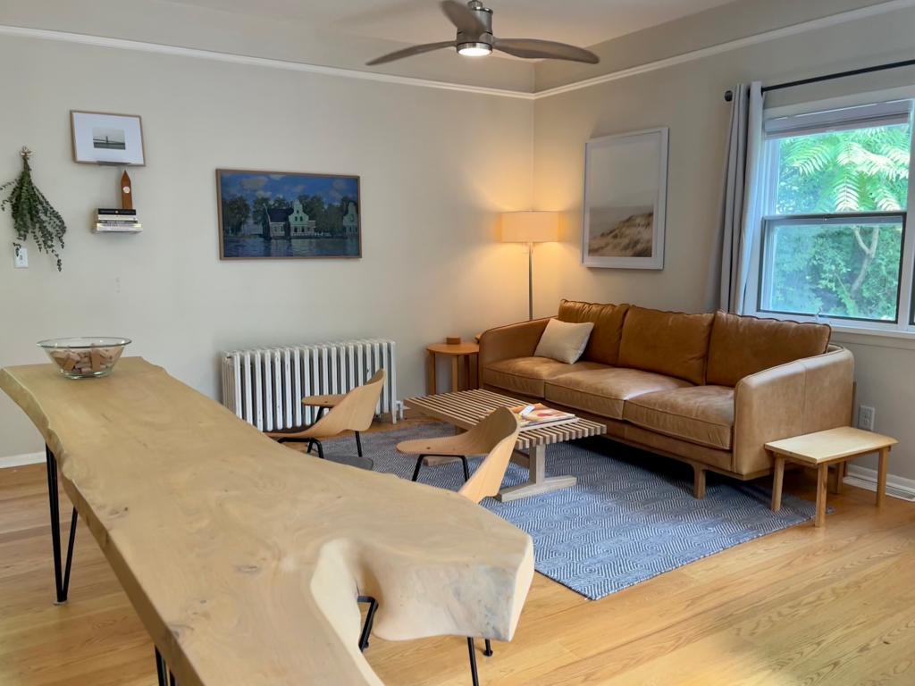 A warm living area features a leather sofa, accompanied by wooden chairs and a low table. A large natural wood dining table enhances the space, while a ceiling fan provides comfort. Light enters through a window dressed with simple curtains, revealing greenery outside.