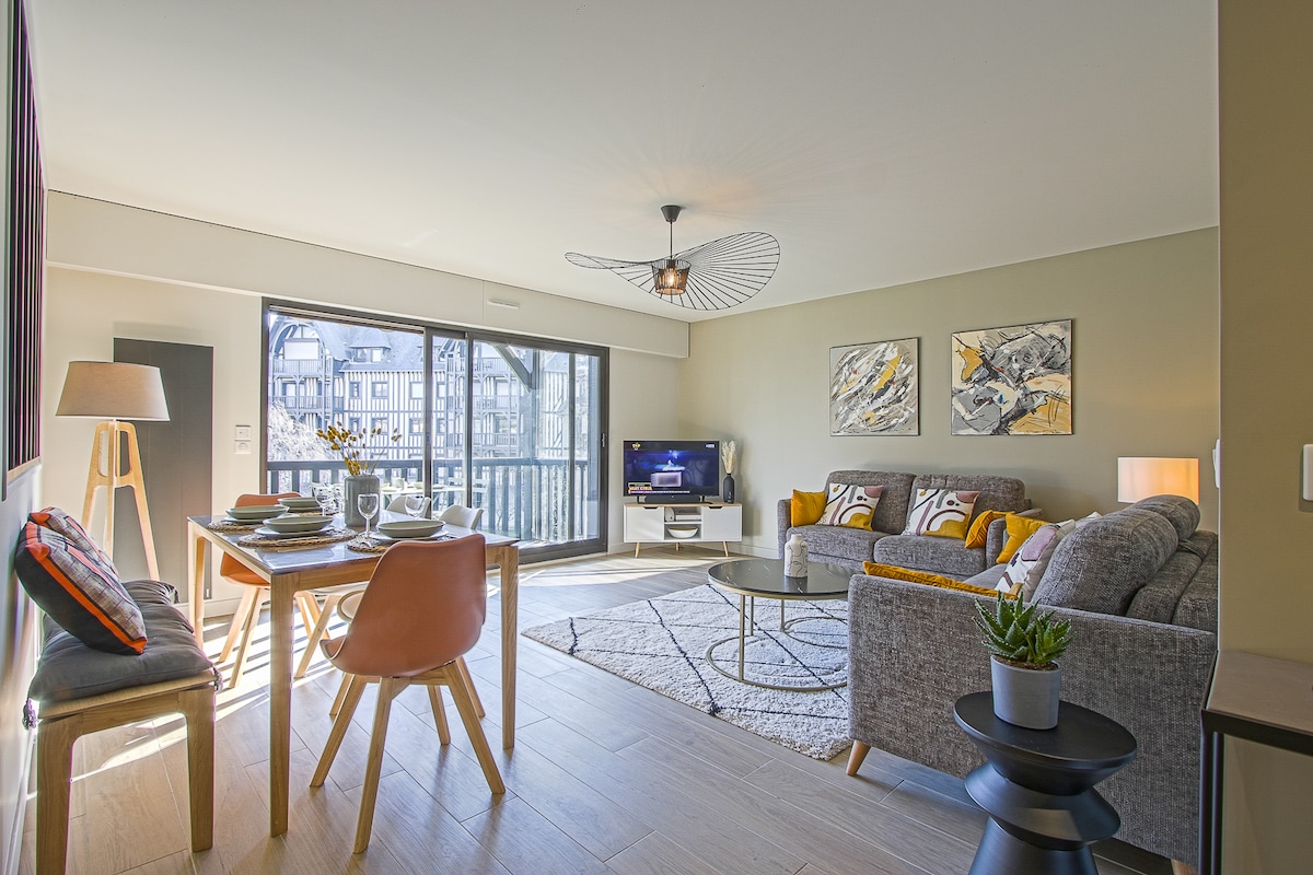 A bright living space features a comfortable sofa and a dining area, complemented by a coffee table. Large glass doors open to a balcony with views of the surroundings, while modern artwork adorns the walls. A television is positioned for entertainment.