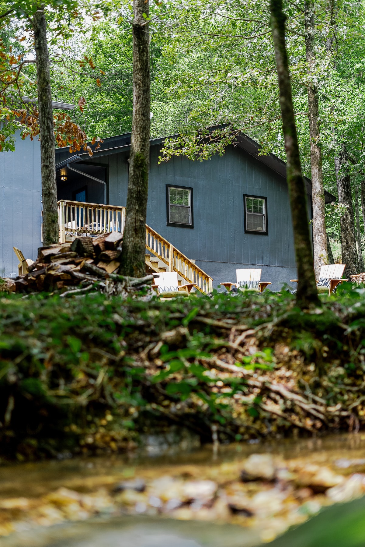 A charming cabin is nestled among lush trees, with a wooden deck offering seating options. A flowing stream is visible in the foreground, surrounded by greenery. The natural setting enhances the feeling of seclusion and tranquility.