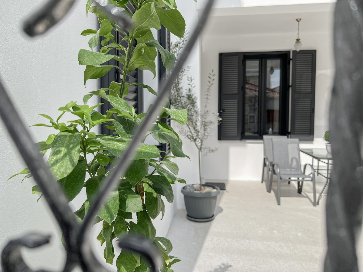 A charming outdoor seating area is framed by greenery and decorative plants. Grey patio furniture is positioned on a light concrete surface, with a small potted tree nearby. Black window shutters complement the white walls of the building, enhancing the peaceful ambiance.