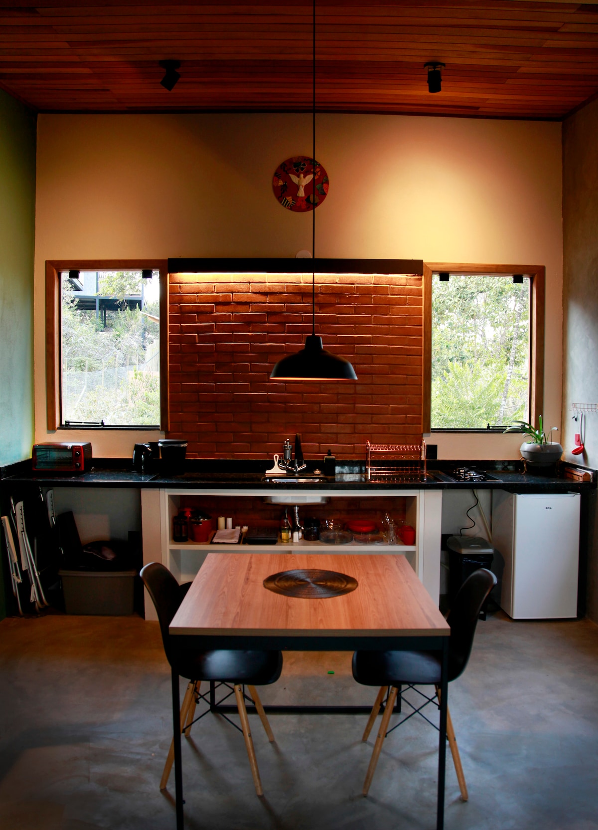 The kitchen features modern black cabinetry with a contrasting wooden accent wall. A circular dining table is paired with black chairs. Two large windows provide natural light and a view of the greenery outside, enhancing the inviting atmosphere of the space.