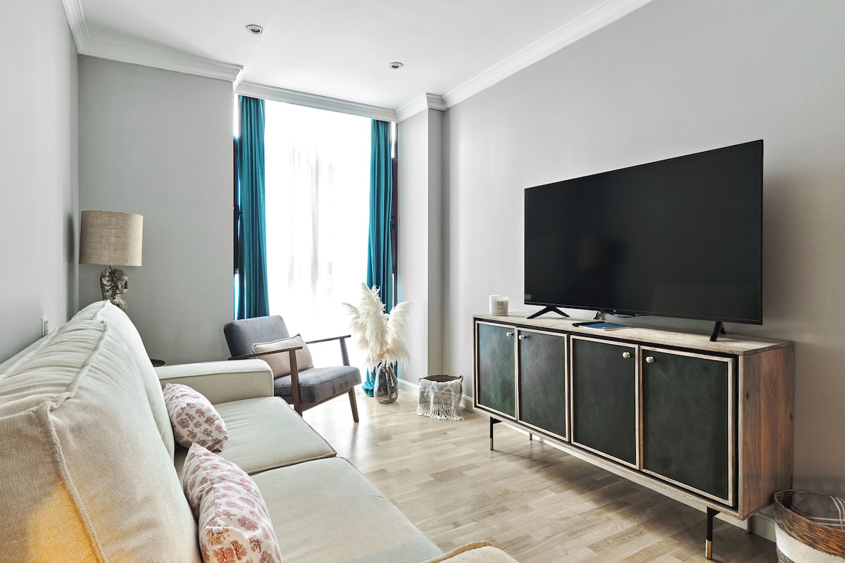 A modern living area features a comfortable sectional sofa with decorative pillows. Natural light filters through two tall windows, adorned with teal curtains. A sleek entertainment unit holds a large television, while a cozy armchair and a decorative plant add to the inviting ambiance.