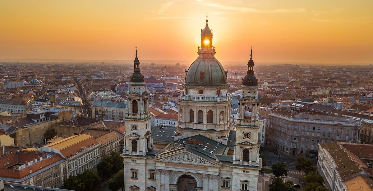 The image captures a breathtaking skyline at sunset, featuring the domed rooftop of a historic building. Towers rise prominently against the evening light, while the cityscape stretches into the distance, highlighted by the warm glow of the setting sun.
