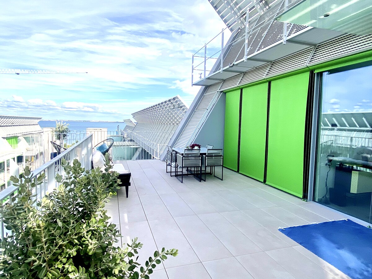 A spacious terrace features a dining table with four chairs, overlooking stunning views of the river. Lush greenery is visible along the edge, complementing the modern architecture of the building. Large sliding doors provide access to the interior, while the sky is bright and clear.