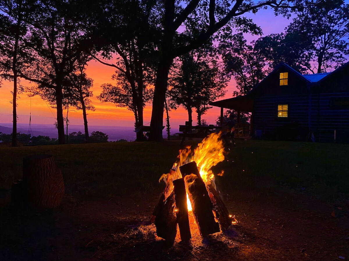 A warm campfire flickers in the foreground, surrounded by a grassy area. The vibrant colors of sunset, including shades of orange and purple, illuminate the sky. In the background, a cabin with windows aglow stands among trees, creating a serene natural setting.