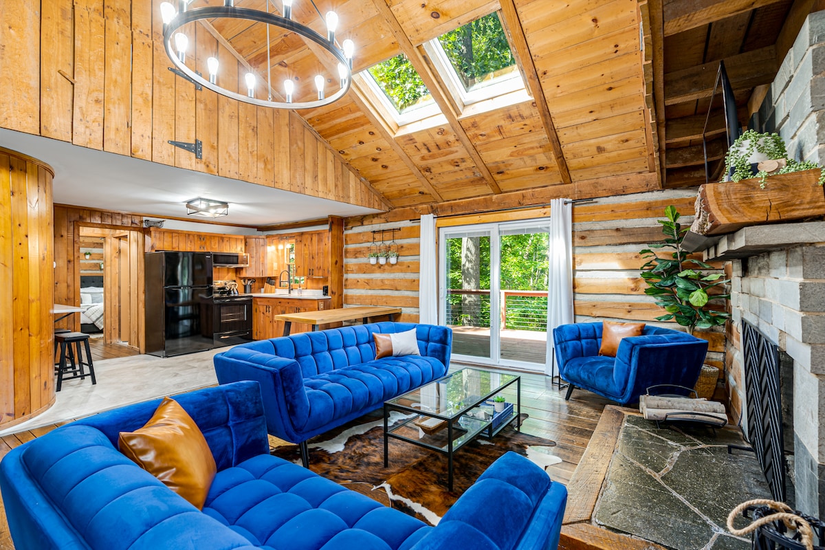 A spacious living area features blue tufted sofas arranged around a glass coffee table. Natural light enters through a skylight and sliding glass doors, which open to the surrounding greenery. Warm wood paneling and a stone fireplace enhance the cabin's rustic charm.