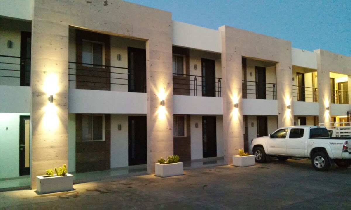 A modern building exterior is shown, featuring a series of well-lit balconies and large windows. A white truck is parked in front, with planters adding a touch of greenery to the entrance area.