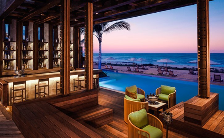 An open-air bar area features warm wooden accents and modern seating. Two green chairs are positioned near a low table, complementing the wooden bar backdrop. Panoramic views of the beach and ocean are visible through the large windows, with soft hues of twilight in the sky.