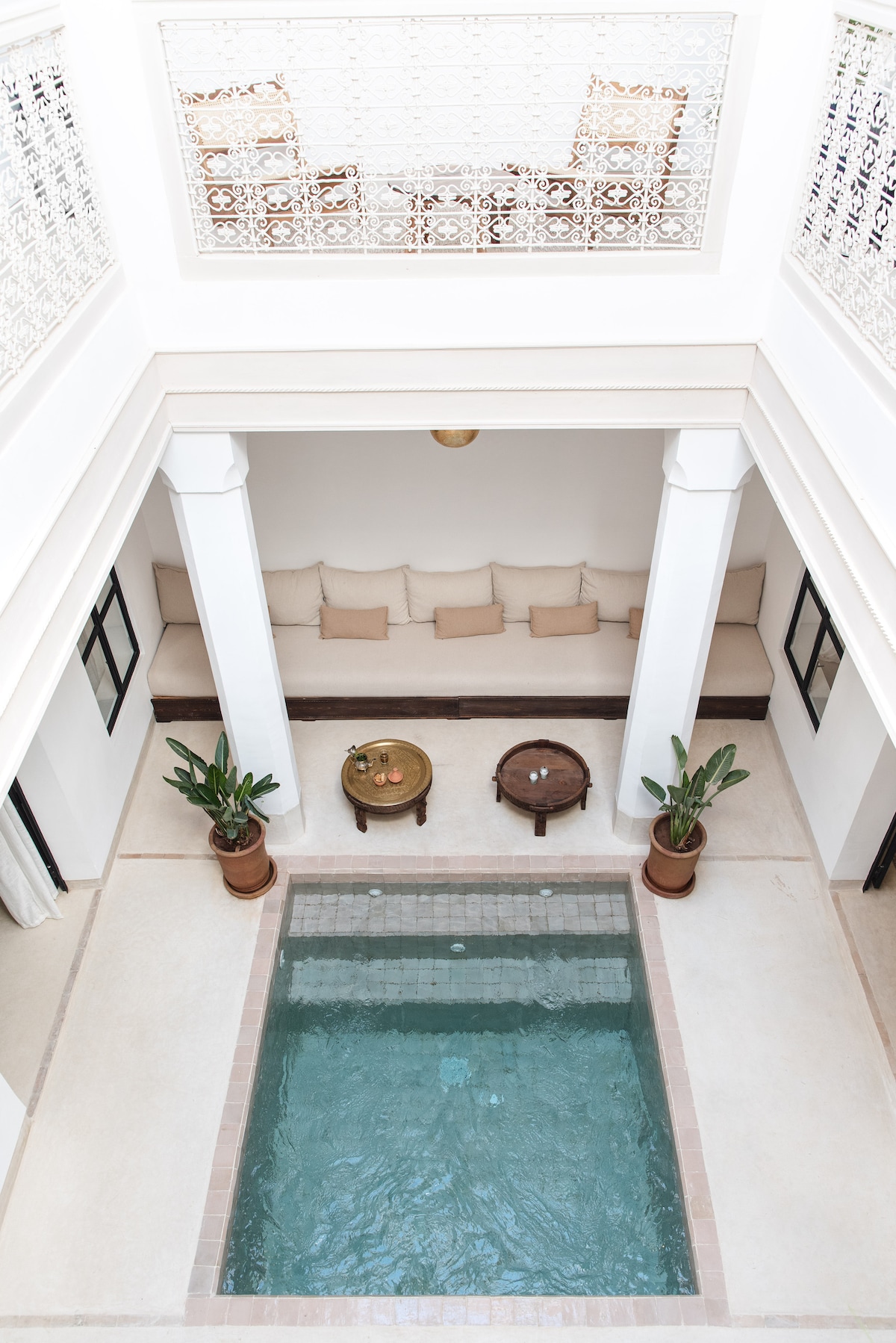 The central courtyard features a heated pool surrounded by earthen-toned tiles. Two potted plants add greenery, while a cushioned seating area is located along the perimeter. Decorative tables complete the inviting space, with light filtered through the upper balcony's intricate latticework.