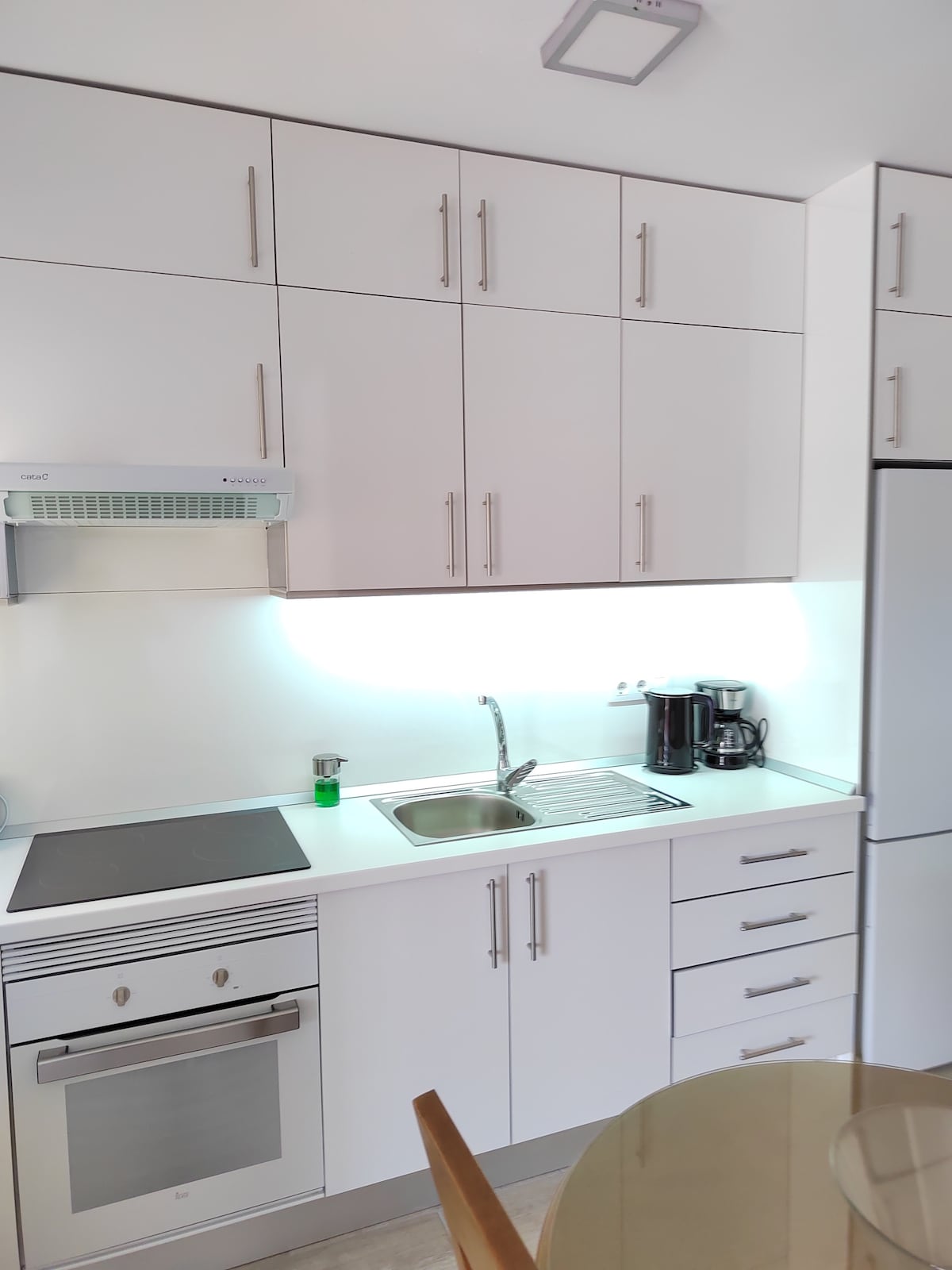 A modern kitchen features sleek white cabinetry, complemented by a metallic stove and built-in oven. A stainless steel sink is installed below an illuminated backsplash. Small kitchen appliances, including a coffee maker, are neatly arranged on the countertop. A circular dining table is visible in the foreground.