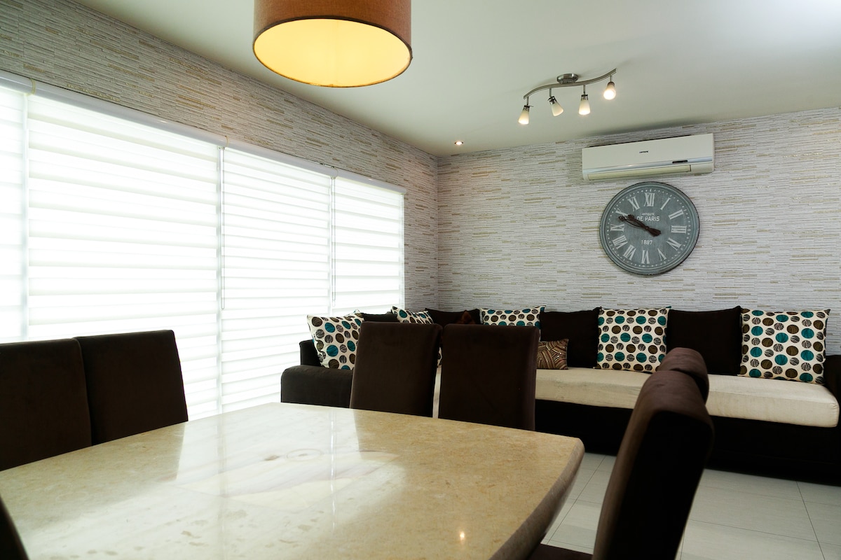 A dining area is visible with a stone-topped table surrounded by chairs. A spacious seating arrangement lines the wall, adorned with cushions featuring a circular pattern. Natural light filters through the window shutters, illuminating the textured walls and a large wall clock.