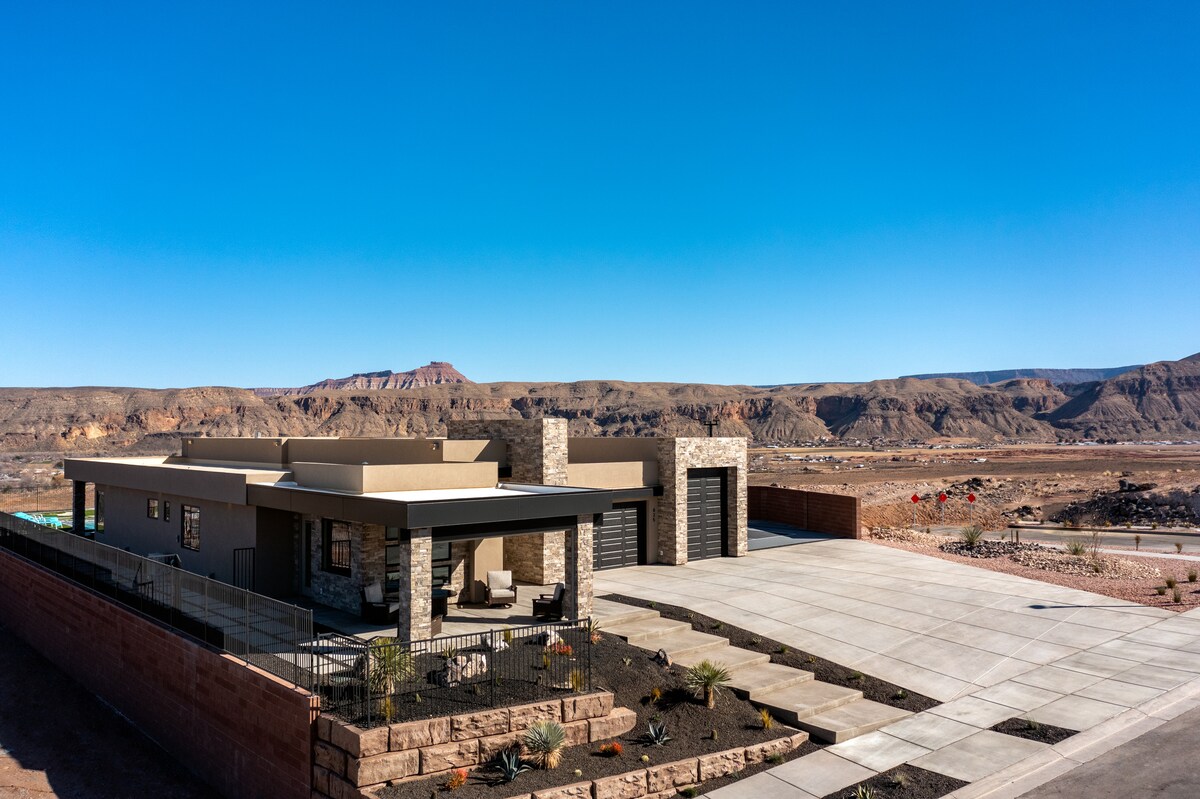 The exterior view captures a modern casita-style guestroom set against a clear blue sky. The structure features a mix of stone and stucco, with a spacious driveway leading to a double garage. Scenic mountainous terrain is visible in the distance.