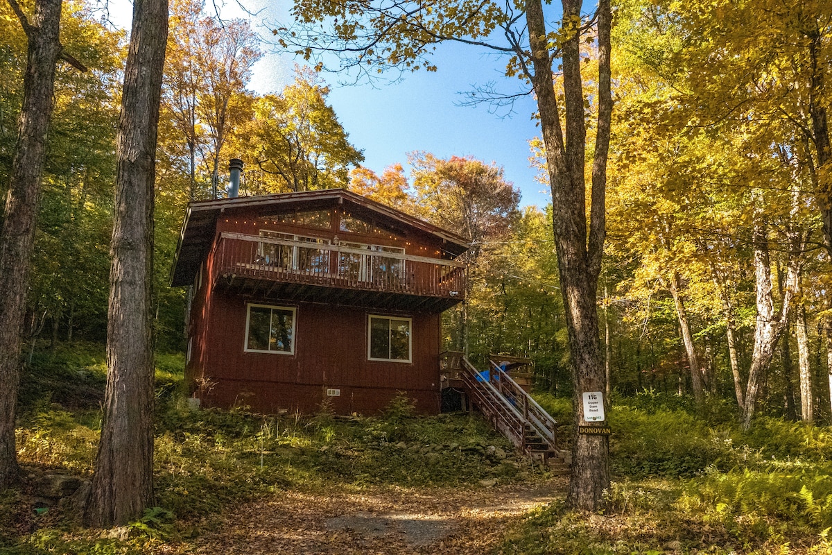 A two-story red cabin is framed by colorful autumn foliage. Wooden steps lead to a front porch with a railing, while large windows allow natural light to illuminate the interior. The surrounding landscape features a variety of trees with leaves in vibrant shades of yellow and orange.