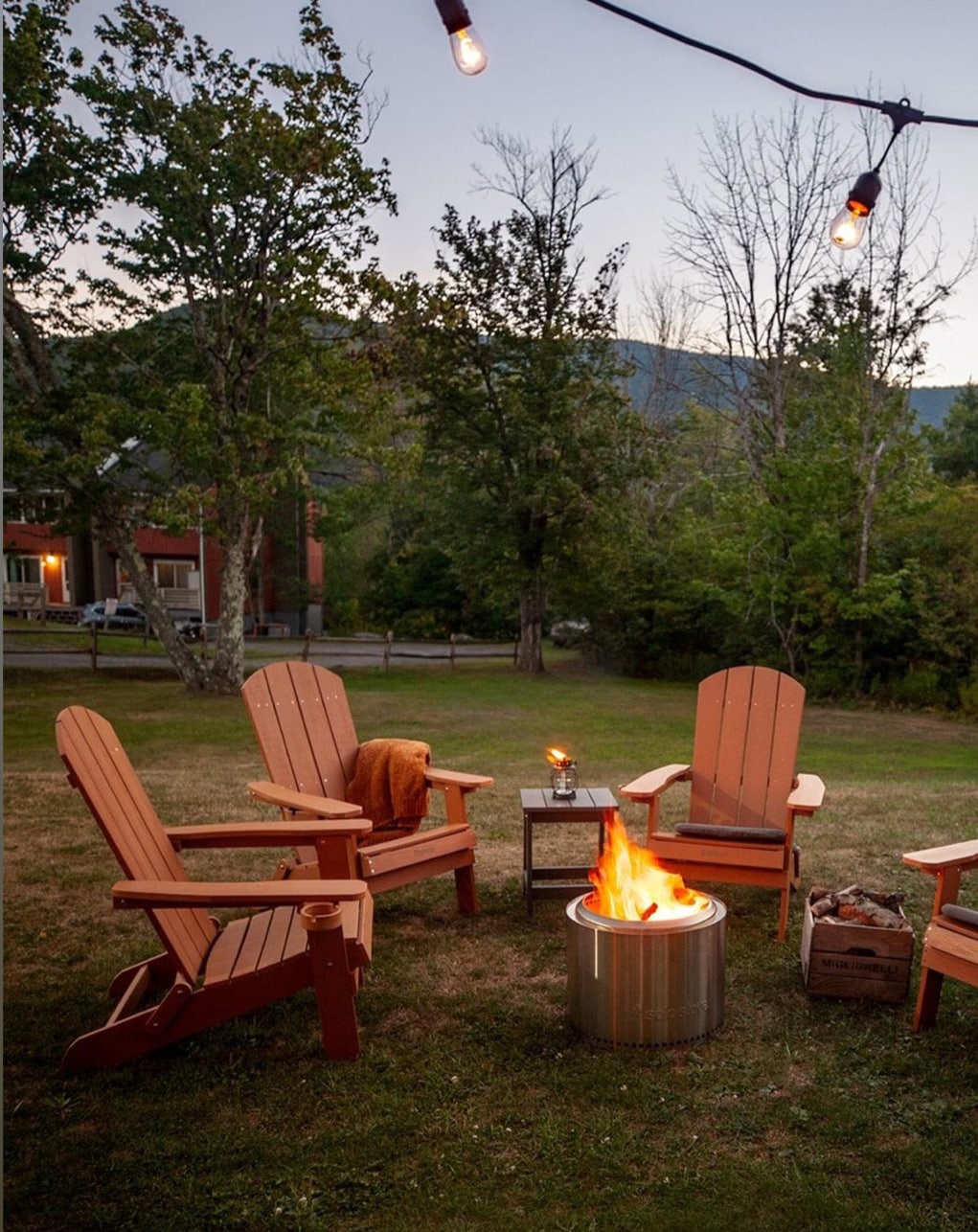 A cozy outdoor seating area features four wooden adirondack chairs arranged around a fire pit. A small table holds a lantern beside the fire, which emits a warm glow. Trees and distant mountains are visible, creating a serene natural setting.