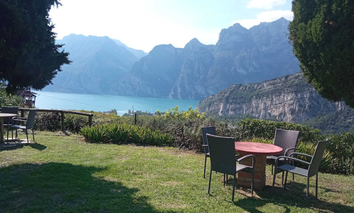 A natural garden area features a round table surrounded by four chairs, set against a backdrop of lush greenery. In the distance, the expansive view reveals Lake Garda and the dramatic mountain scenery, enhancing the sense of tranquility.