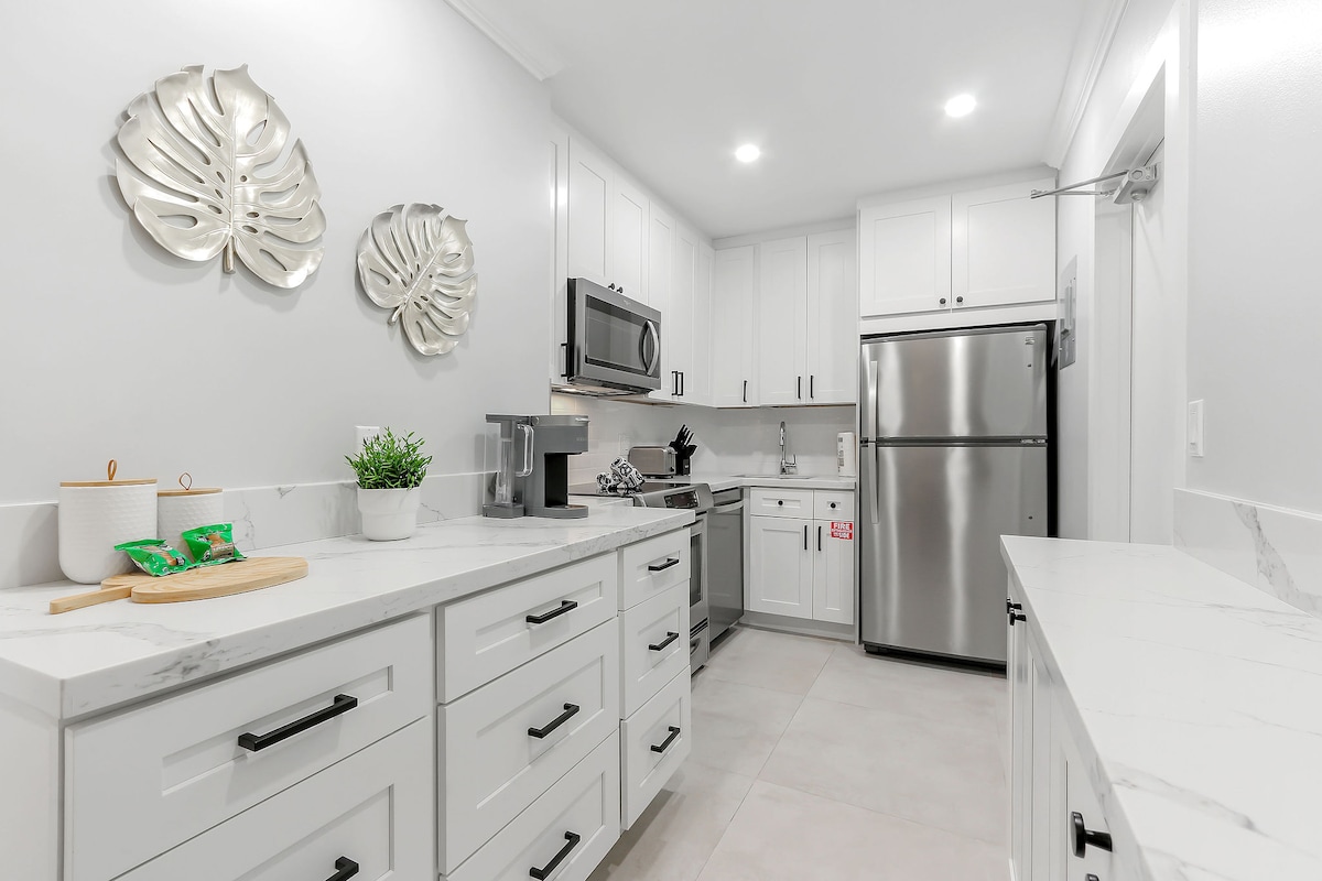 The kitchen showcases modern cabinetry in white, complemented by marble countertops. Stainless steel appliances, including a refrigerator and microwave, are present. Decorative wall art resembling large leaves adds a subtle touch. Ample counter space is available for meal preparation and dining.