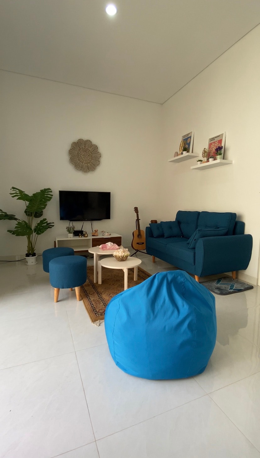 A bright living area features a blue sofa and matching accent chairs. A small coffee table sits in the center, accompanied by a decorative rug. A plant adds a touch of greenery, while a television and guitar are visible on a shelf.