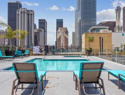 A rooftop pool area is depicted with clear water and surrounding lounge chairs in a vibrant turquoise color. The skyline of downtown Los Angeles serves as a backdrop, showcasing a mix of modern buildings and historic architecture under a bright blue sky.