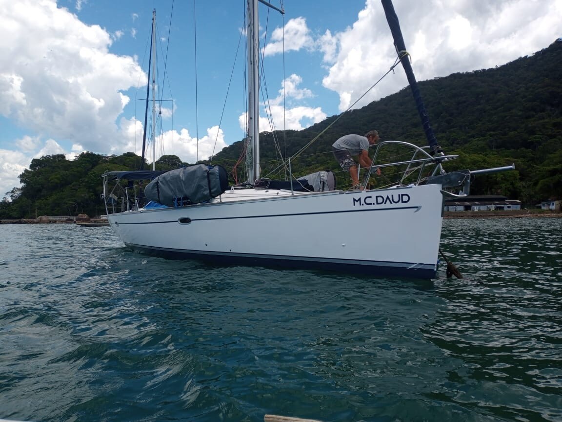 A sailboat is seen anchored in clear water, surrounded by lush greenery. The boat features a white hull and is equipped with a canopy, partially covering the deck. A figure can be seen preparing equipment on the vessel, with a backdrop of rolling hills and blue sky.