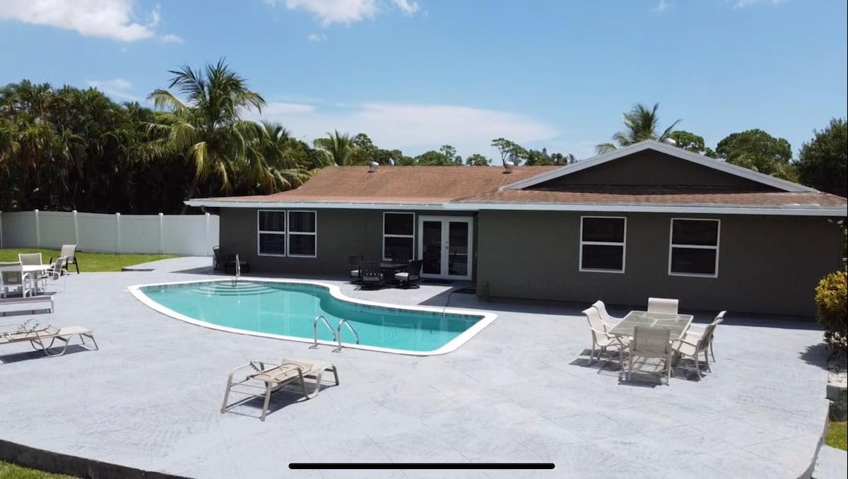 A spacious outdoor area features a clear swimming pool surrounded by a textured patio. Several lounge chairs and a dining set are positioned around the pool, with tropical palm trees and a privacy fence visible in the background.