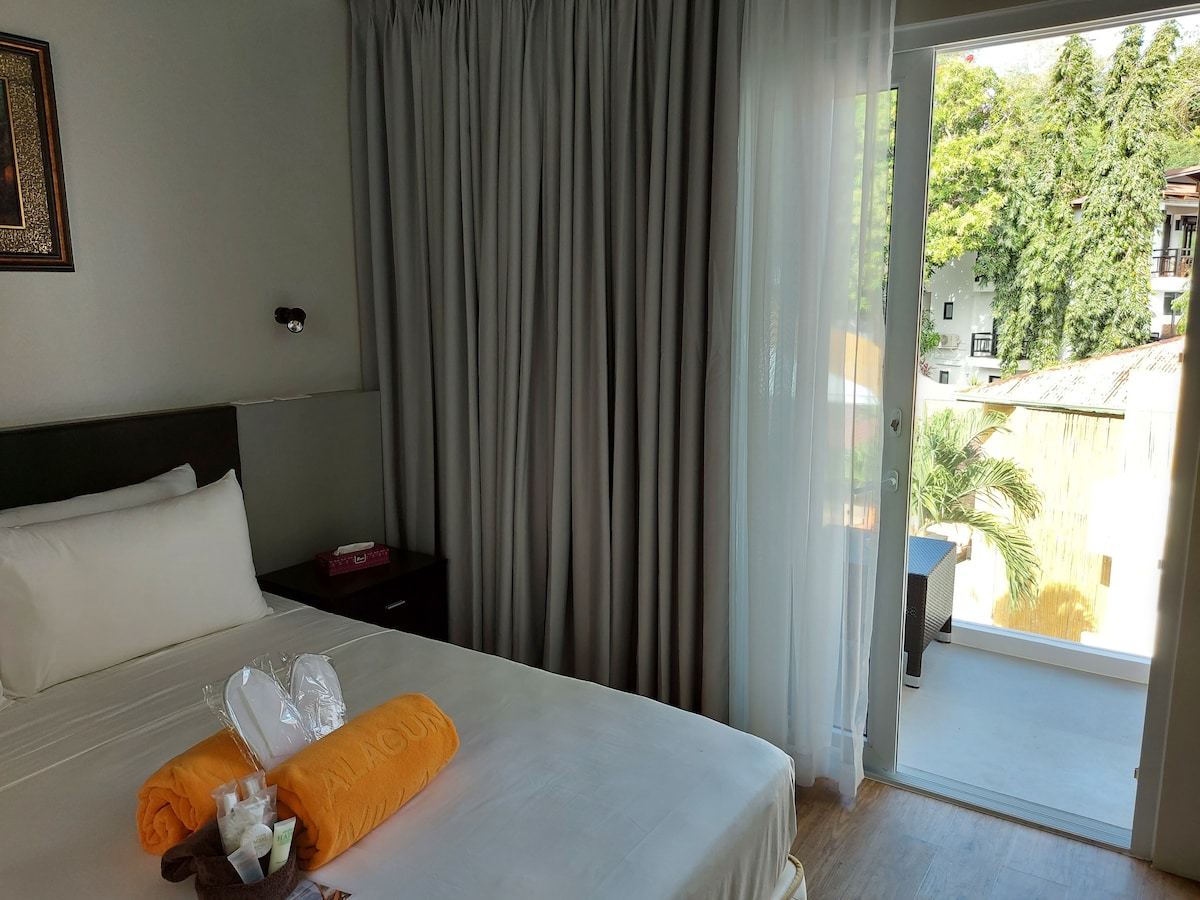 A bright bedroom is presented with a spacious bed dressed in crisp white linens and accented by vibrant orange towels. Floor-to-ceiling curtains frame a large glass door that opens to a private balcony, revealing lush greenery just outside.