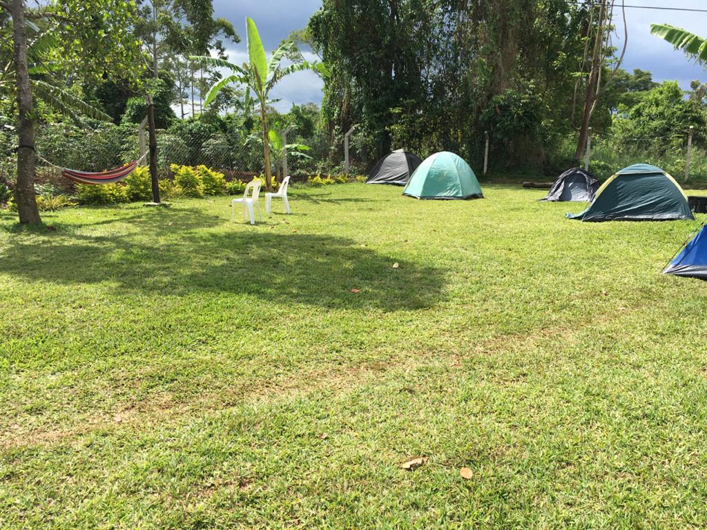 A grassy campsite is visible, showcasing several tents arranged on the well-maintained lawn. In the foreground, two white chairs are positioned, offering a place to relax. A hammock is draped between two trees, surrounded by lush greenery and scattered shade.