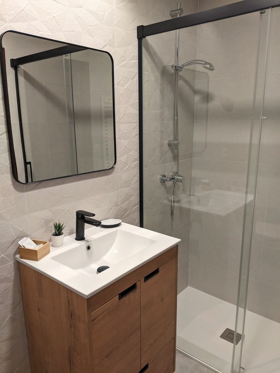 A modern bathroom features a glass shower enclosure and a sleek vanity with a white sink. The decor includes neutral-toned wall tiles and a large mirror. A small plant adds a touch of greenery to the minimalist space.