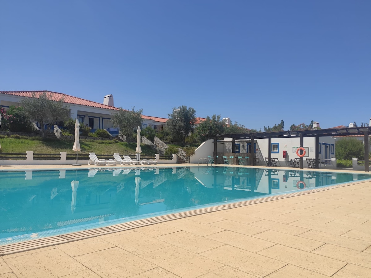 A large outdoor swimming pool reflects the clear blue sky, surrounded by well-maintained greenery. Several lounge chairs are arranged alongside the pool, and shaded areas are provided by umbrellas. Nearby, a residential building is visible, completing the tranquil setting.