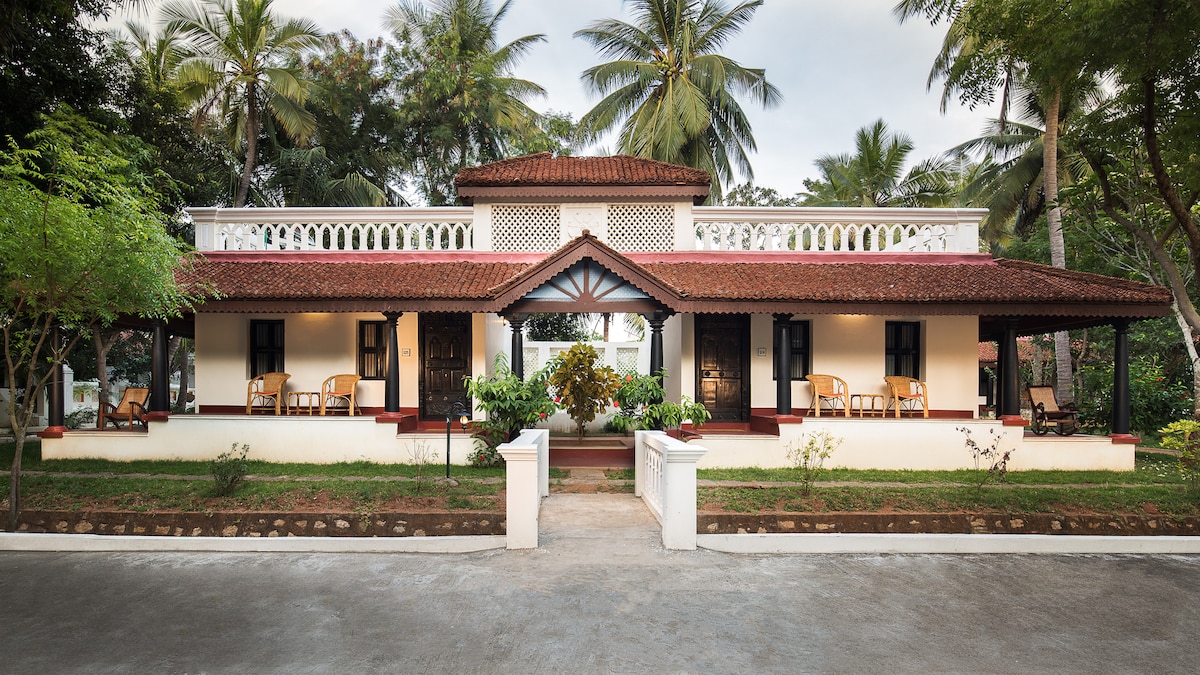 A charming cottage is framed by lush greenery and palm trees, featuring a traditional tiled roof and a welcoming façade. Verandahs are adorned with wooden chairs, inviting guests to enjoy the serene surroundings. The well-maintained garden adds to the tranquility of the property.