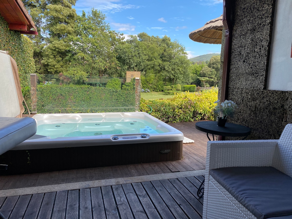 A private jacuzzi is positioned on a wooden deck, surrounded by lush greenery. The serene outdoor space includes a small table and a comfortable chair, inviting relaxation while enjoying views of the well-maintained lawn and trees in the distance.