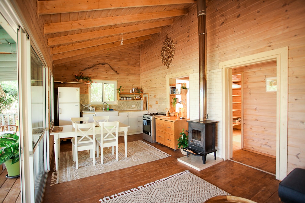 A warm wooden interior features an open kitchen and dining area with a large table and white chairs. A stove adds rustic charm, while light floods in through multiple windows, illuminating the natural wood paneling and enhancing the cozy atmosphere.