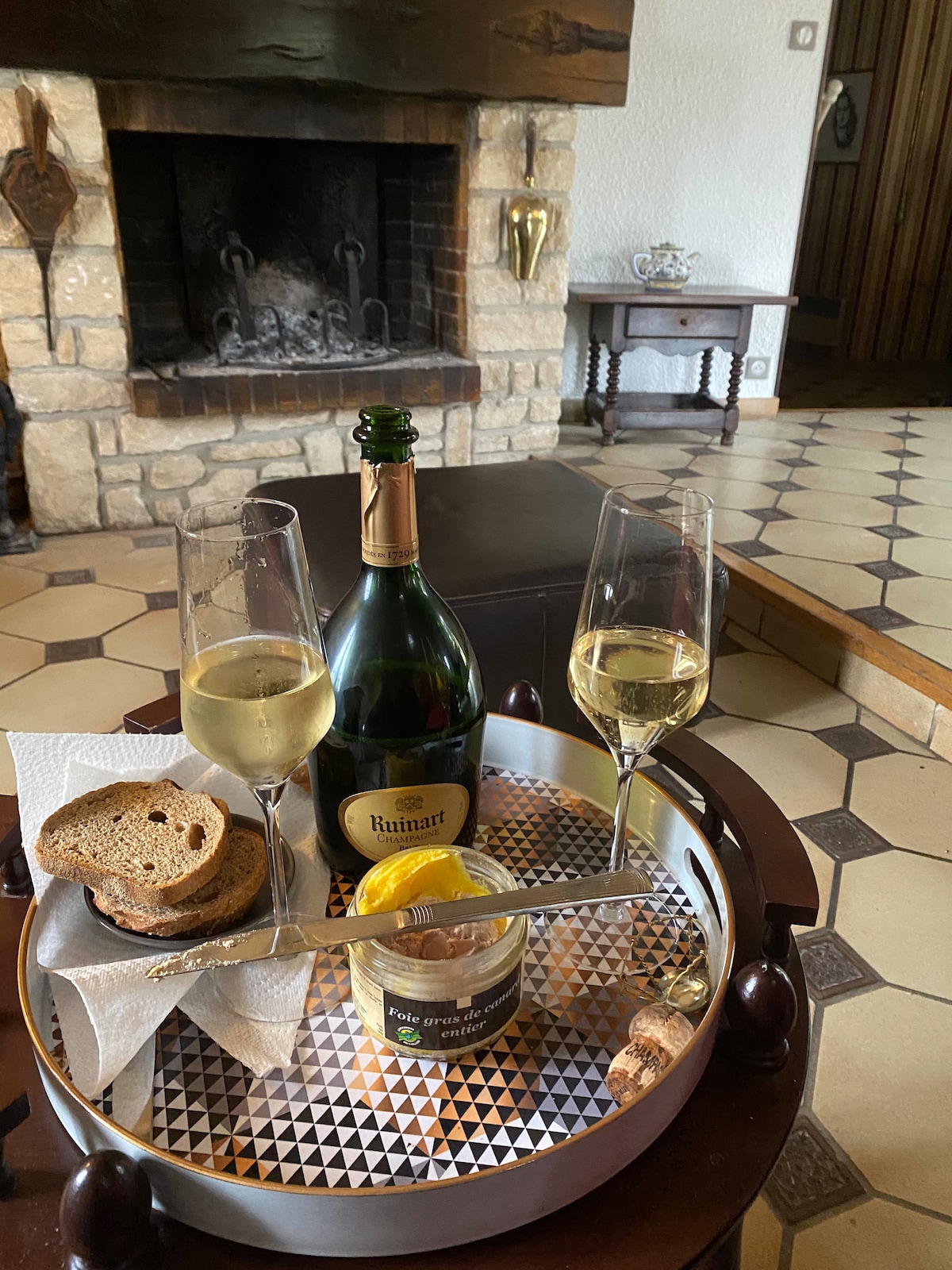 A wooden tray presents two glasses of bubbly champagne alongside a bottle. Slices of bread and a small jar of spread are arranged neatly, complemented by a lemon slice. In the background, a stone fireplace adds warmth to the inviting interior.