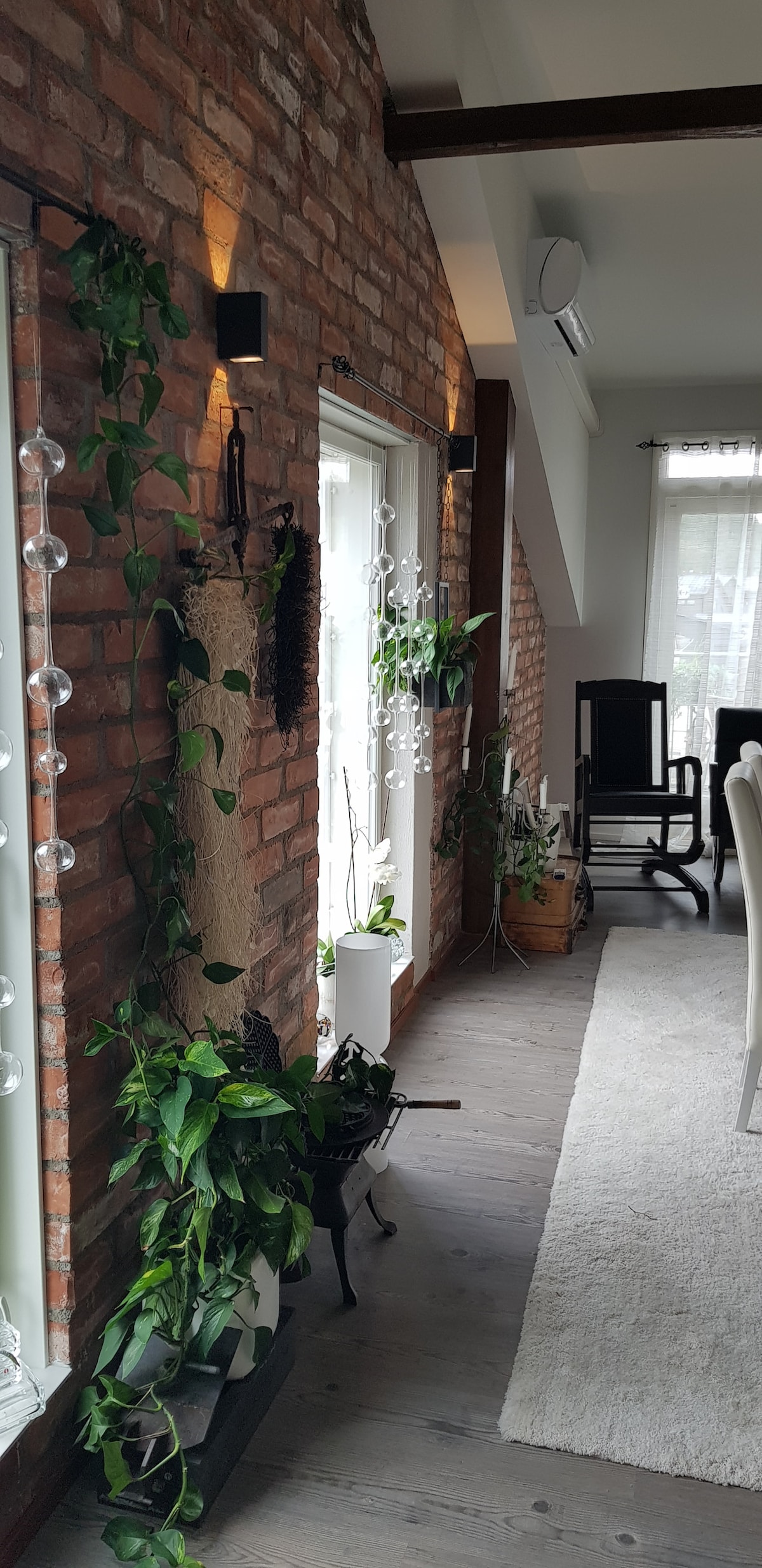 Exposed brick walls create a rustic charm in the living space. Natural light filters through large windows, with indoor plants adorning the sides. A light area rug adds texture to the hardwood floor, complementing the room's welcoming design.
