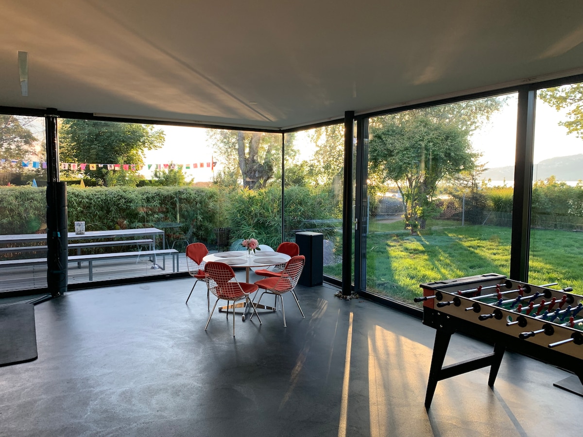 A spacious living area is surrounded by floor-to-ceiling glass walls, providing views of the garden and greenery. An oval dining table is surrounded by red chairs, and a table soccer game is positioned nearby, allowing for both dining and leisure activities.