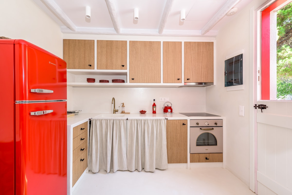 A modern kitchen features a bright red refrigerator, light wood cabinetry, and a white countertop. A white curtain drapes over the sink area, and a stove with an oven is positioned beside a flat-screen microwave. Natural light enters through the open door.