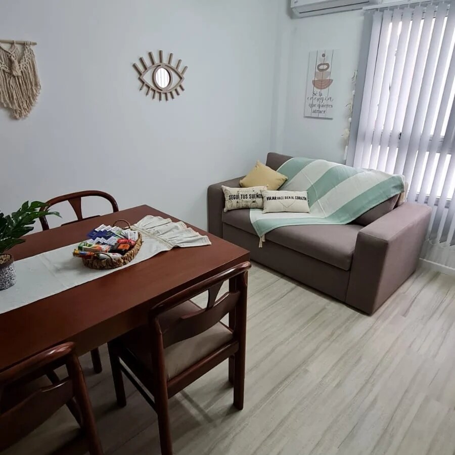 A neutral-toned living area is presented with a compact dining table and four wooden chairs. A slipcovered sofa is positioned nearby, adorned with a light throw. Decorative wall art and a round mirror add subtle character, while natural light filters through vertical blinds.