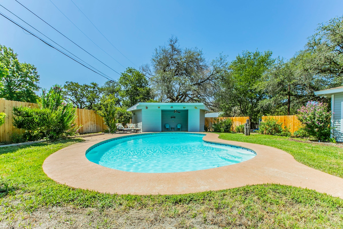 A smooth, rounded swimming pool is set in a spacious, private backyard, surrounded by lush greenery and flowering bushes. A shaded seating area is visible behind the pool, providing a comfortable spot to relax. The clear blue water invites leisurely enjoyment under the sunny sky.