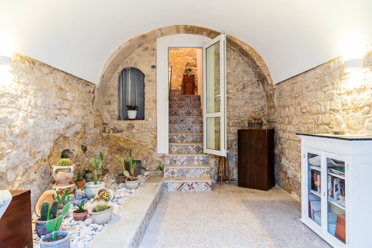 An archway leads into a welcoming entry space with rustic stone walls. Decorative potted plants are positioned along the floor, while a staircase with patterned tiles ascends to the main area. Natural light filters through a nearby window, enhancing the serene atmosphere.