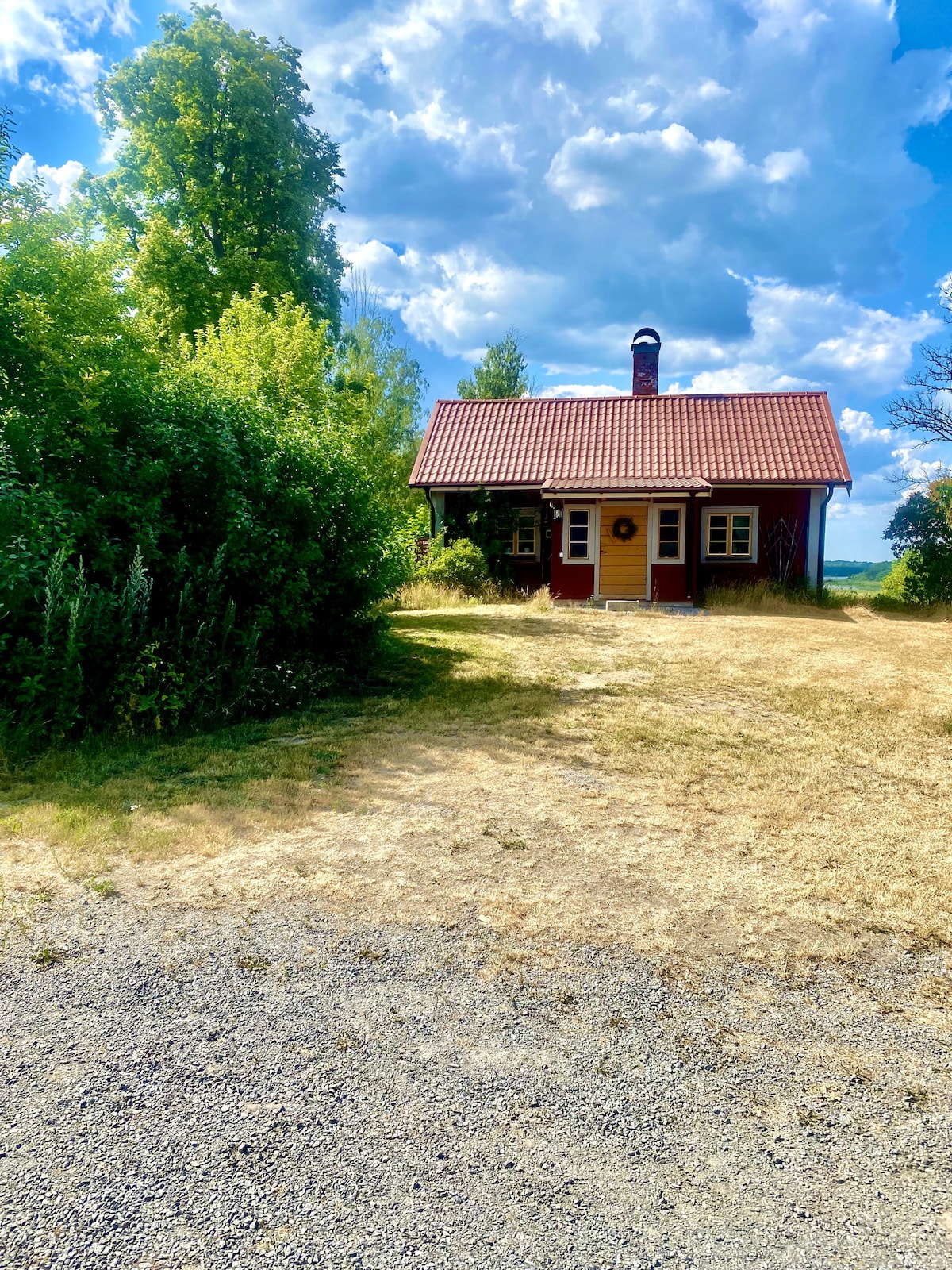 A charming 1800s cottage is positioned amidst lush greenery, featuring a red exterior with white accents and a brown tiled roof. A welcoming front porch is visible, complemented by surrounding grassy areas and trees under a blue sky with scattered clouds.