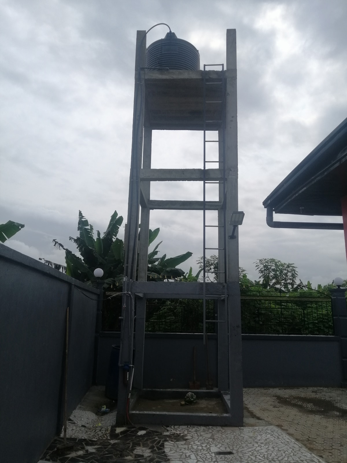 A tall, concrete water tower is seen against a cloudy sky. The structure is elevated and includes a ladder for access. Surrounding plants provide greenery, while a patterned stone pathway is visible at the base.