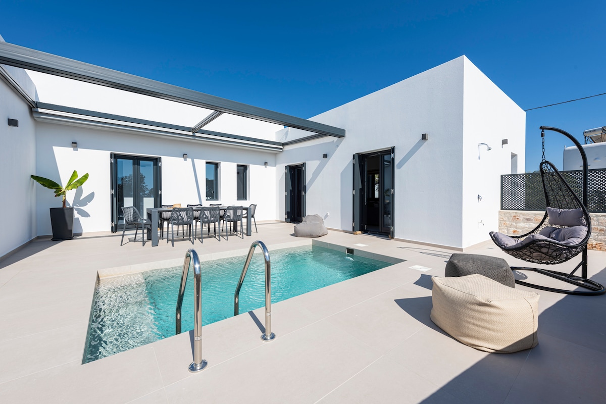 An outdoor area features a swimming pool framed by a spacious sun terrace. A dining table with several chairs is positioned beneath a covered section, while loungers and bean bags add comfort. Clear skies create a bright atmosphere, enhancing the villa's modern architecture.