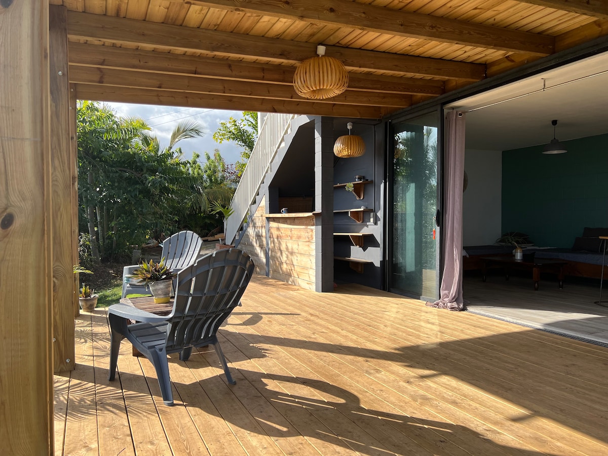 A covered wooden terrace features two comfortable chairs positioned on a warm wooden floor. Lush greenery surrounds the area, enhancing the outdoor space. Glass doors lead into the interior, where natural light is highlighted by decorative light fixtures above.