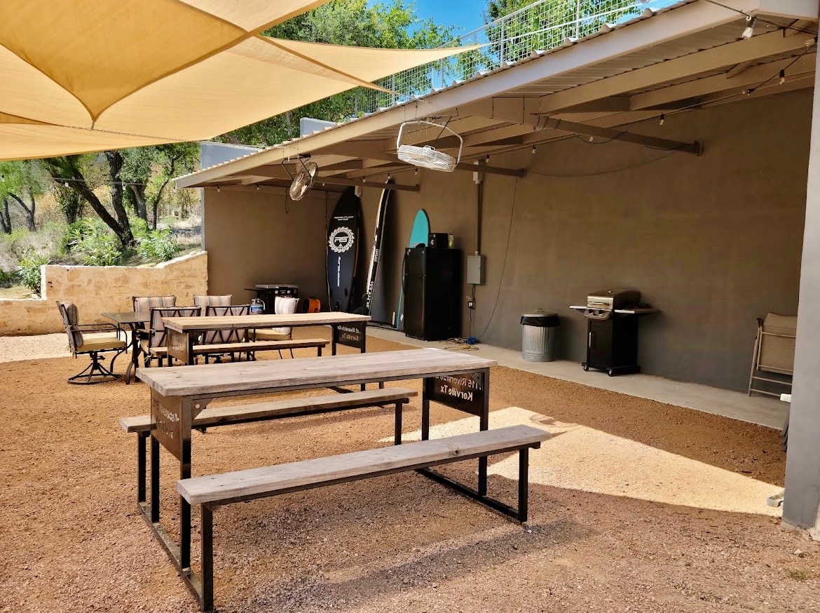 An outdoor patio area features several picnic tables arranged for communal seating. A striped canvas awning provides shade overhead. Grilling equipment and a refrigerator are visible in the background, alongside surfboards. The ground is covered with gravel, enhancing the rustic charm of the space.