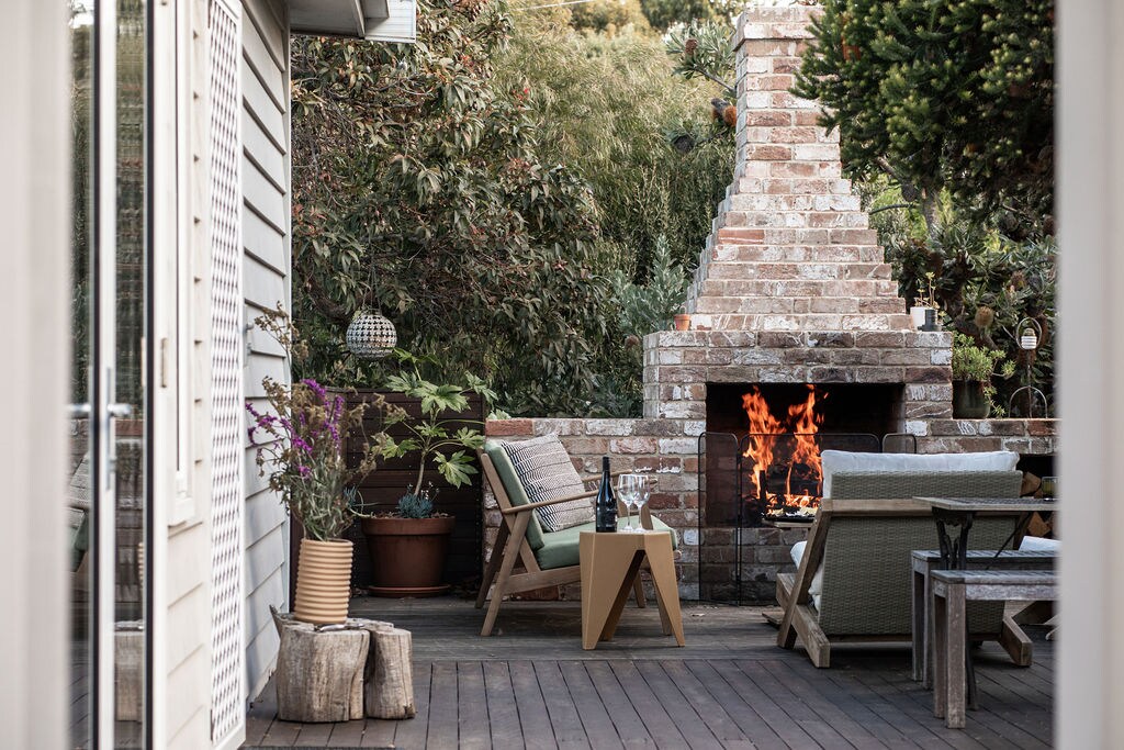 An outdoor living area is seen through an open doorway, featuring a cozy seating arrangement around a brick outdoor fireplace. The fire flickers warmly, surrounded by potted plants. A small table is set with a wine bottle, inviting relaxation and social gatherings.