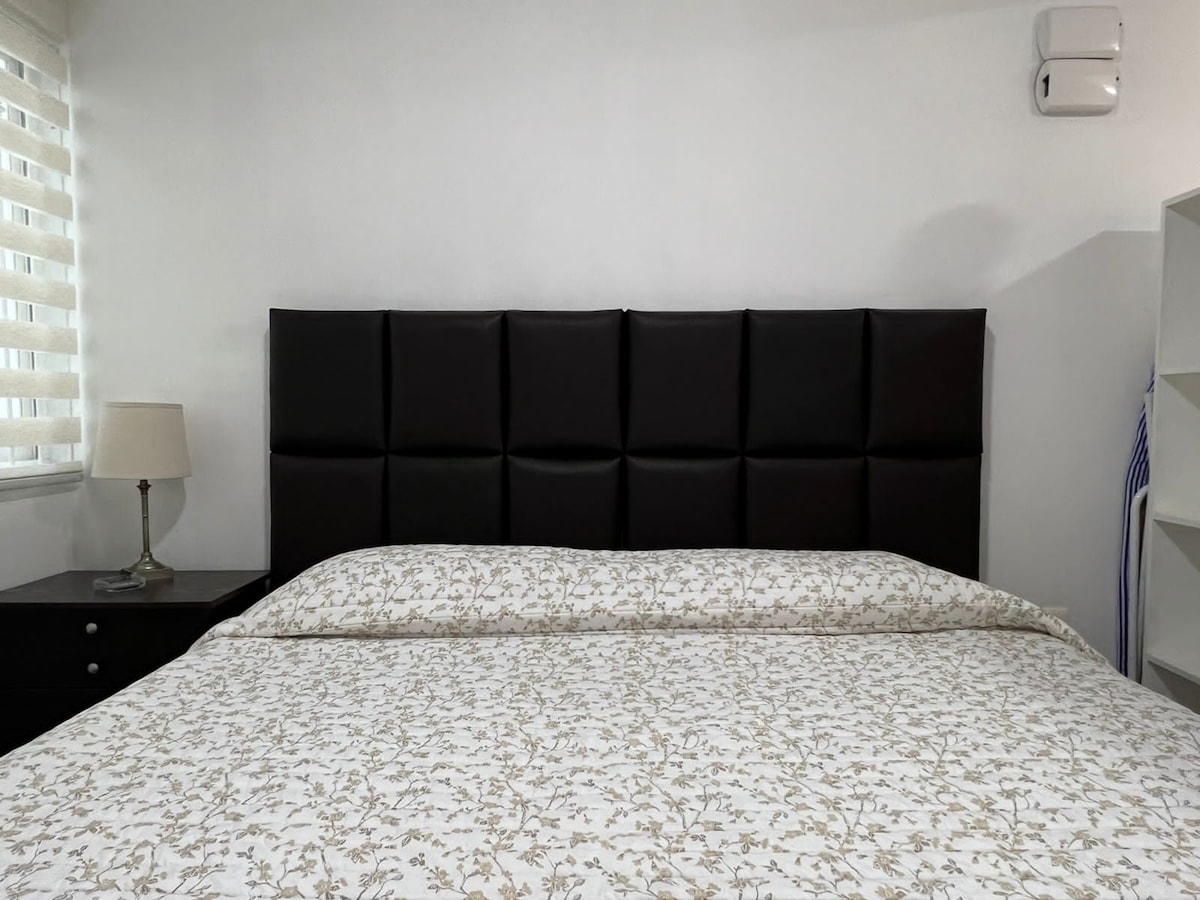 The sleeping area features a king-size bed with a stitched black headboard. A light-colored floral-patterned bedspread covers the bed. A bedside lamp is placed on a nightstand, and a bookshelf is present in the background, enhancing the room's functionality.