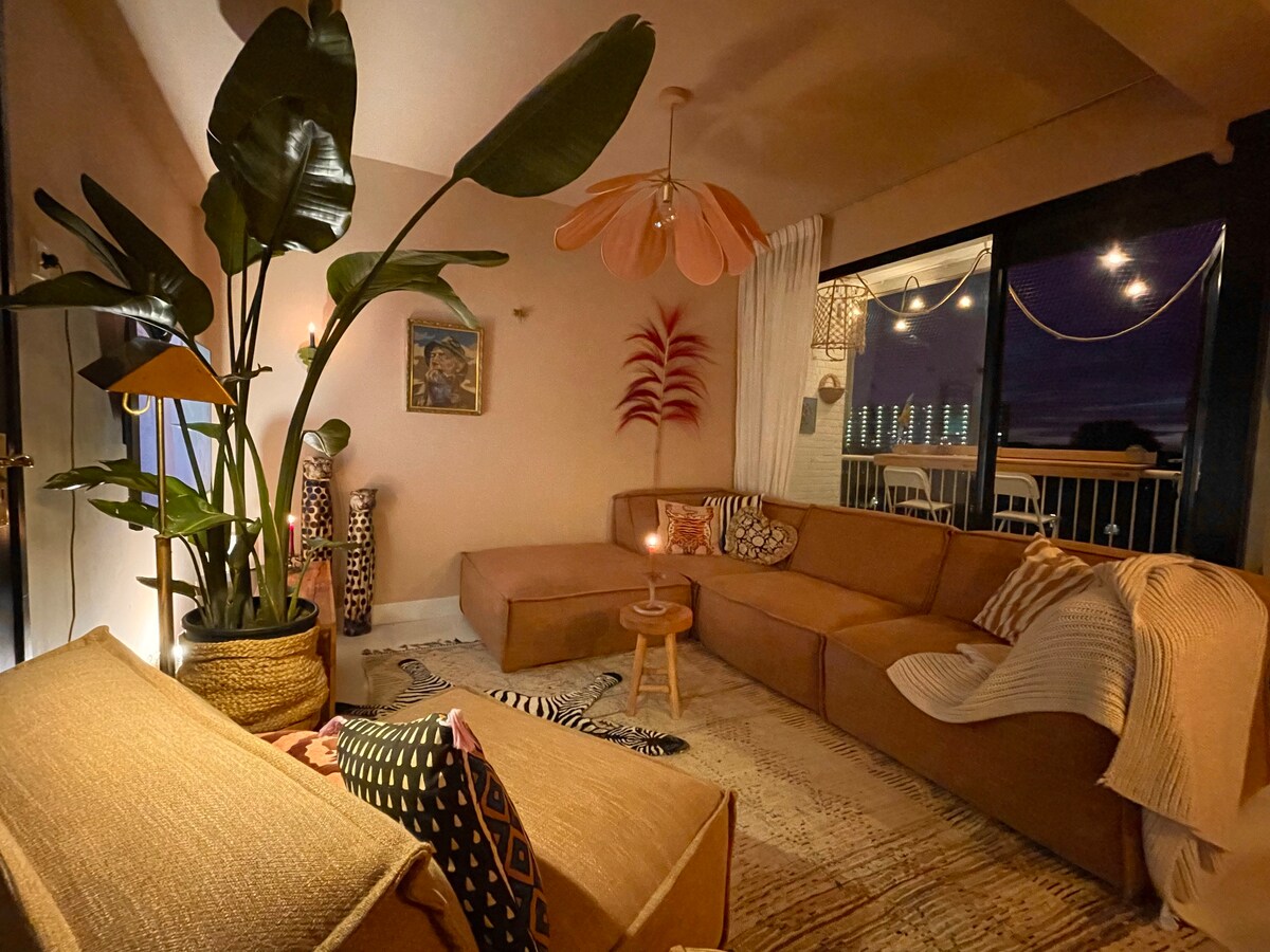 A cozy living area is highlighted by a large sectional sofa, surrounded by indoor plants. Soft lighting emanates from a table lamp, and decorative pillows add comfort. The space opens up to a balcony, providing a view of the outdoors and enhancing the overall ambiance.