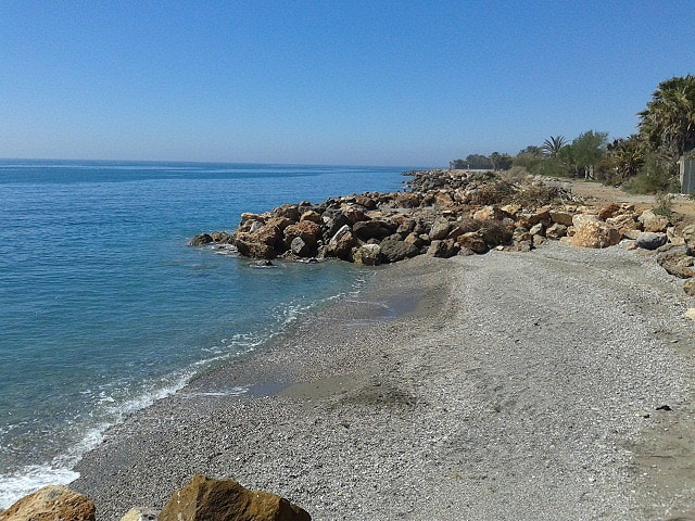 A serene beach scene features a gently sloping shoreline lined with smooth pebbles and coarse sand. Rocky formations stand along the water's edge, with the calm sea extending into the distance. Clear blue skies complement the tranquil atmosphere.